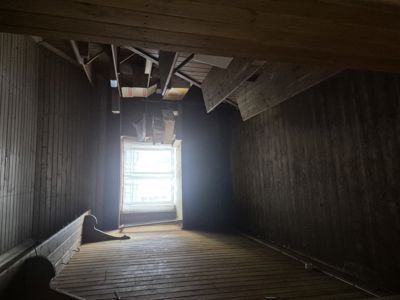 Fotografi av et rom under renovering eller i en rå tilstand, med synlig trepanel på vegger og gulv samt eksponerte takbjelker.