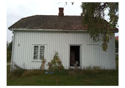 Fotografi av en hvit, trepanelert bygning sett fra fronten. Bygningen har et tak med taktegl, en røykledning og en åpen dør.