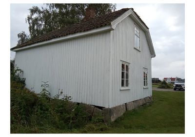 Fotografi av en hvit, tømmerbygning sett fra siden og bak. Bygningen har et skrått tak, en røykpipe og står på betongfundament.