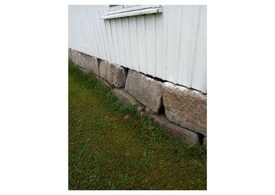 Fotografi av bygningens nedre del, som viser hvit panelvegg, vinduslist og en steinmur som fundament.