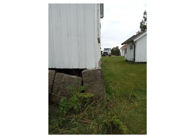 Fotografi som viser nederste del av en hvit bygning med steinfundament, samt en annen hvit bygning i bakgrunnen og en bil.