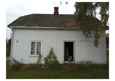 Fotografi av en hvit, trepanelert bygning sett fra fronten. Bygningen har et tak av teglfliser, en rødmalt kamin, et vindu og en dør. Det er også synlig vegetasjon og en del av en annen bygning i bakgrunnen.