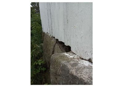 Fotografi av en bygning som viser en skade eller sprekke i fundamentet der det hvite tregulvet møter steinmuren.