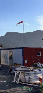 Fotografi av en byggeplass med et rødt og hvitt containerbygg, en flaggstang med norsk flagg, og materialer på bakken.