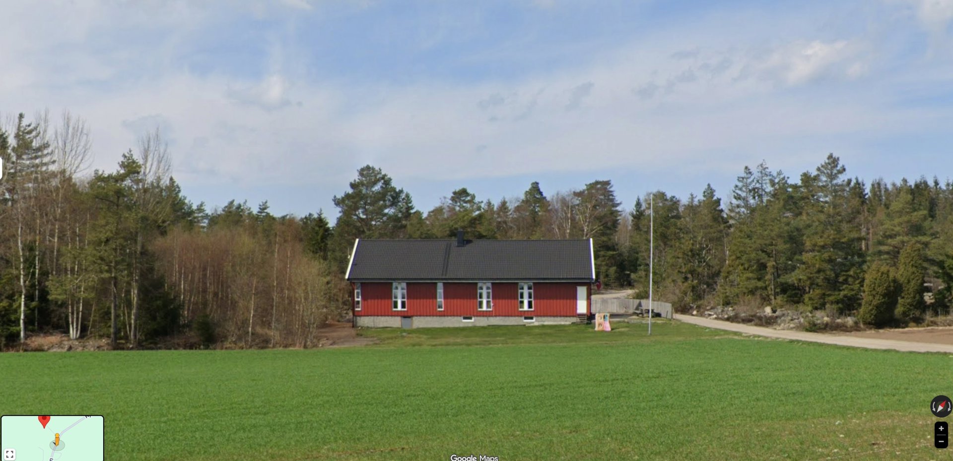 Fotografi av en rød enebolig med grå tak sett fra et felt mot en skogbakke.