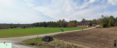 Fotografi av en eksisterende bygning (rød hytte) sett fra en vei, med omgivende natur og landbruk.