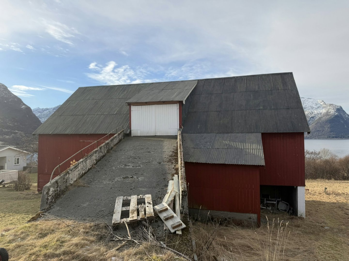 Fotografi av en rød skure eller uthus med hvit garasjeport, sett fra fronten mot bakgrunn av fjell og vann.