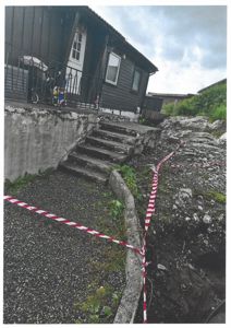 Fotografi av en eksisterende bygning (tømmerhus) sett fra siden, med trapp ned til en grusflate og en utgravd del av bakken. Bildet viser også en advarselstape.