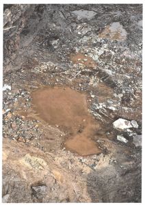 Fotografi av en våt, mudderete og steinete overflate, sannsynligvis en graving eller et område med vannlekkasje.