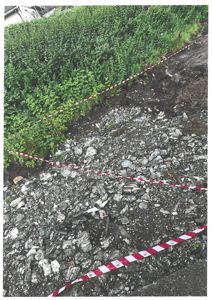 Fotografi av en steinete bakke eller terreng med en rød og hvit advarselstape som markerer et område. I bakgrunnen ser man busker og en del av en bygning.