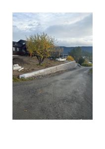 Fotografi av en byggeplass eller eiendom med en nybygd betongmur, en vei, trær og en eksisterende hytte i bakgrunnen.