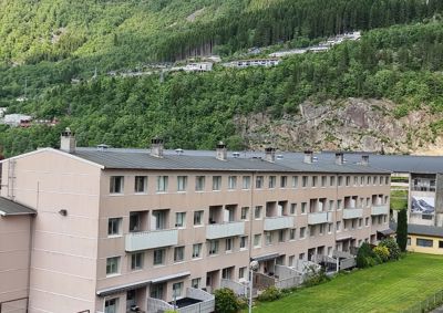 Fotografi av en fleretasjes bygning (sannsynligvis et boligblokk) sett fra siden, med bakgrunn av en skogkledde fjellside.