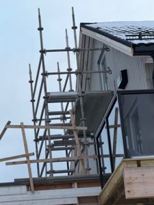 Fotografi av en byggeplass med en nybygd husfasade, tak og en stor stige (scaffolding) ved siden av.