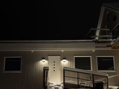 Fotografi av en bygning sett fra utsiden om natten. Bildet viser fasaden med vinduer, en dør, utvendig belysning og en trapp opp til en veranda. Det er snø på taket og trappen.