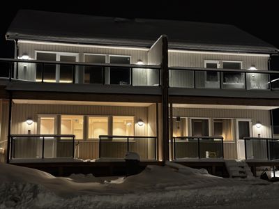 Fotografi av en toetasjes bolig eller leilighetsbygg sett fra gaten. Bygningen er belyst om natten, og det er mye snø på taket, balkongene og i forgrunnen.