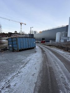 Fotografi av en byggeplass med en stor grå industribygning, en blå søppelcontainer, en kran og en snødekket vei.