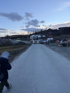 Fotografi av et byggplass med en lastebil som kjører inn på en ny asfaltert vei, med en person i forgrunnen.