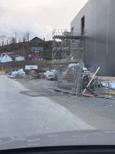 Fotografi av en byggeplass med en stor, mørk fasade på høyre side, en byggeplatt (scaffolding) og materialer i forgrunnen.