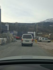 Fotografi tatt fra bil som viser et byggplassområde med materialer, kjøretøy og en bygning i bakgrunnen.