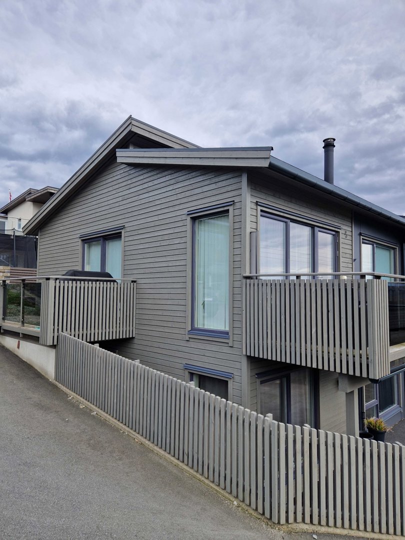 Fotografi av en moderne, grå beleggset bolig sett fra gaten. Bildet viser fasaden, balkonger, vinduer og en hekk.