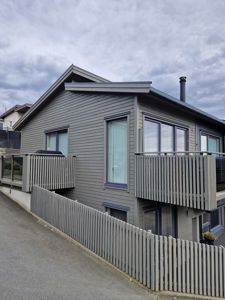 Fotografi av en moderne, grå beleggset bolig sett fra gaten. Bildet viser fasaden, balkonger, vinduer og en hekk.