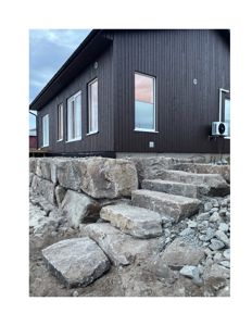 Fotografi av en bygning med mørke fasadepaneler, vinduer og en luftkondisjonsenhet, sett fra bakkenivå mot en steinmur.