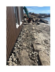 Fotografi av en byggeplass ved kysten, der en grønt gaffeltruck jobber med å rydde eller grave i stein og grus langs en bygning med brun fasade.