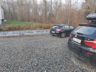 Fotografi av en parkeringsplass med to biler, en hekk og en del av et hus.