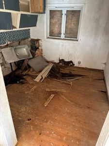 Fotografi av en skadet eller demontert kjøkkenrom med opprisset gulv, skadet vegg og en vask.