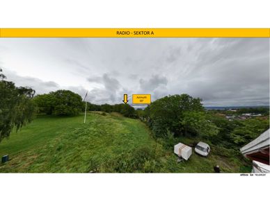 Fotografi av et utsiktspunkt med vegetasjon, en flaggstang og en bygning i forgrunnen. Bildet inneholder også overlagte tekster og en pil som indikerer retning.