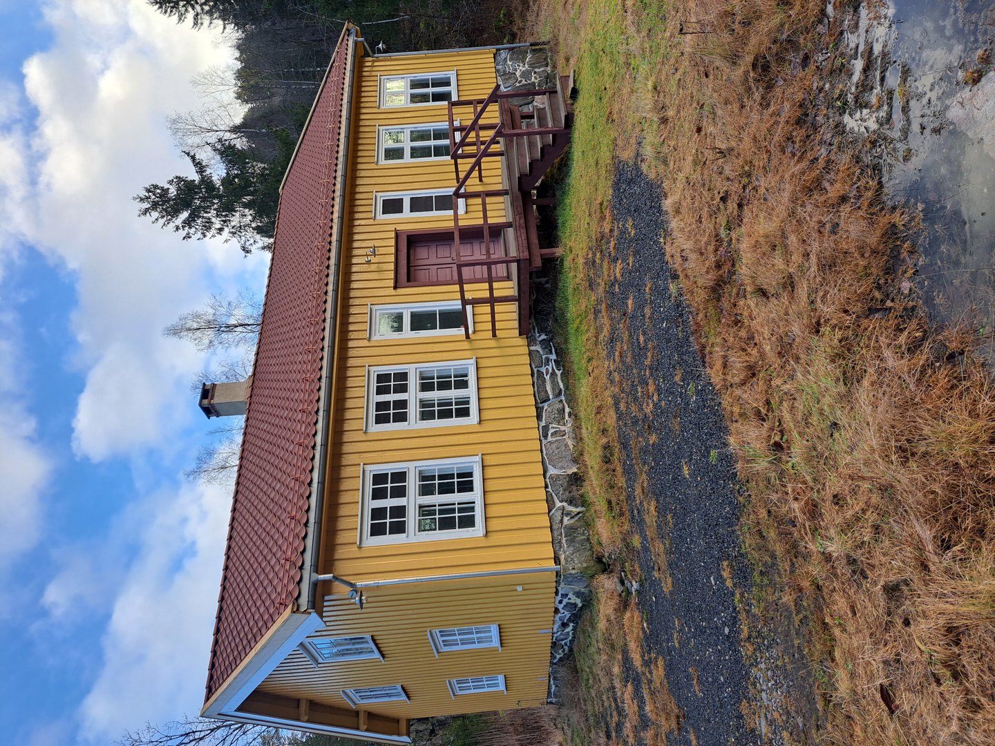 Fotografi av en gul farget bygning med rødt tak sett fra siden, med trapp opp til inngang.