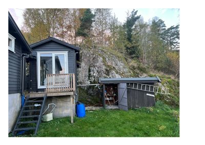 Fotografi av en grå hytte med en liten veranda, en uthus i bakgrunnen og en steil, skogkledt bakke.