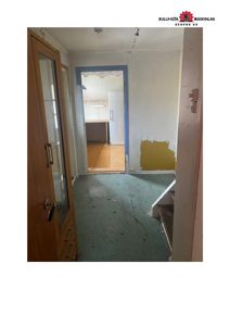 Fotografi av en tom, delvis renoveringsklar leilighet eller bolig. Bildet viser en gang med gulvbelegg, en dør til kjøkken, og en trapp opp til etasjen over.