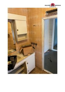 Fotografi av et bad i en bygning under renovering eller ombygging. Bildet viser en vask, speilskap, fliser og en døråpning til et annet rom.