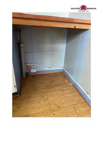 Fotografi av en innendørs hjørne i en bygning under ombygging eller renovering. Bildet viser en gulvet av sponplater, en hvit rørledning langs veggen, en elektrisk uttak, og en overhengende trekonstruksjon.