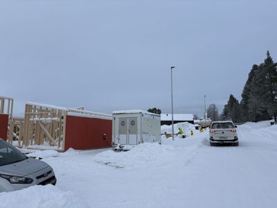 Fotografi av et byggplass i vinterforhold med snø. Bildet viser en delvis oppført trebygning (rammeverk), en hvit container, en bil og en person i gul vest.