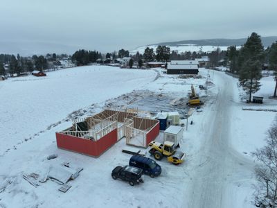 Fotografi av en byggeplass i vinterlandskap. Bildet viser et hus under oppføring med oppstilt tømmervegger, samt byggemaskiner og kjøretøy på snødekt tomt.