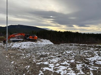 Fotografi av et byggefelt med gravemaskin, snø og skog i bakgrunnen.