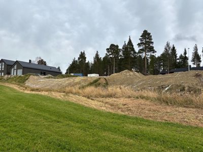 Fotografi av et byggefelt med grønt gress i forgrunnen, en utgravd eller oppdyrket jordhøyde i midten, og en mørk bygning i bakgrunnen mot en skog.