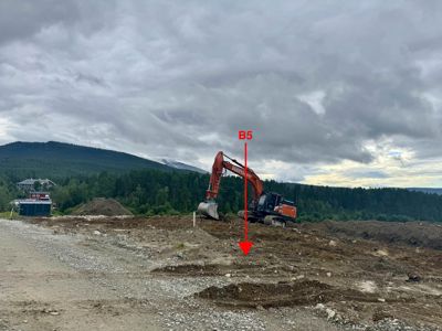 Fotografi av et byggefelt med en graverende maskin, en grusvei og en bakgrunn med skog og fjell.