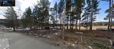 Et panoramafoto tatt fra Google Street View som viser et tomt, skogkledt område med en vei i forgrunnen og noen spredte bygninger i bakgrunnen.