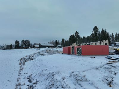 Fotografi av en byggeplass i vinterforhold. Bildet viser en nybygget husstruktur med opprisset tak og røde fasadevegger, omgitt av snø og skog.