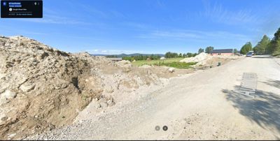 Fotografi av et byggefelt med utgravet jord, en grusvei og en bygning i bakgrunnen.