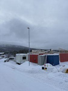 Fotografi av et byggplass under vinterforhold med snø, en delvis ferdigstilt bygning med rødt panel og trekonstruksjon, samt container og hyttebil.