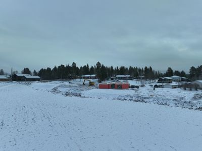 Fotografi av et byggefelt i vinterlandskap med snødekte bakker, skog og flere boliger i bakgrunnen. I midten er det en byggeplass med en gul graver og en nybygget struktur med røde paneler.