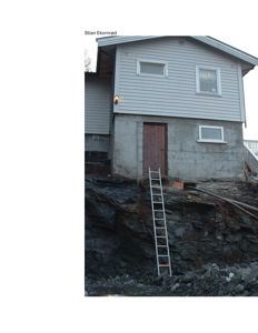 Fotografi av en bygning med grå fasade og betongfundament, sett fra siden med en trapp og steinvegg i forgrunnen.