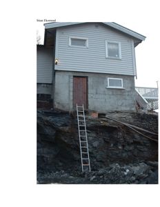 Fotografi av en toetasjes bolig med grå fasadeplater og betongfundament, sett fra siden med en steil fjellside i forgrunnen.