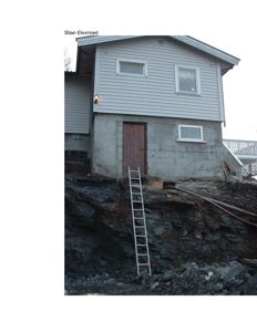 Fotografi av en toetasjes bolig med grå fasade, betongfundament og en trapp opp til etasjen. Bildet viser også en steil fjellside under bygningen.