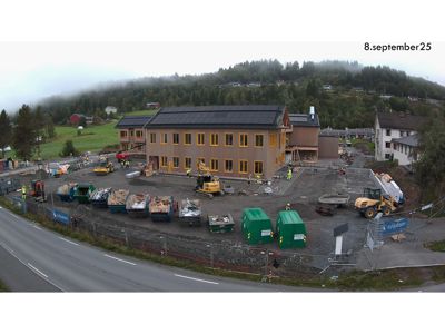 Fotografi av et byggprosjekt under utførelse, med en ferdig fasade på hovedbygningen, maskiner, materialer og arbeidere på tomten.