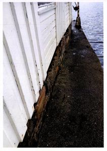 Fotografi som viser en nærbilde av en bygning med hvite panelvegger og en steinmur ved vannkanten.